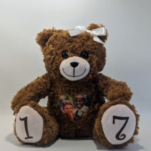 An image of a brown bear with a white bow, a photo of a couple on it's heart, and the number "1" on the left paw, and the number "7' on the right paw. It sits against a plain white background.