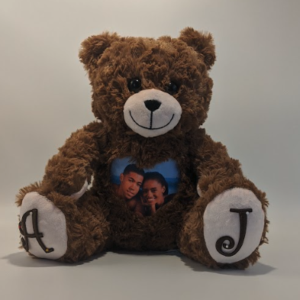 A photo of a mocha brown teddy bear with a photo on its heart and paw engravings, with "A" on the left paw, and "J" on the right paw. It sits against a plain white background.
